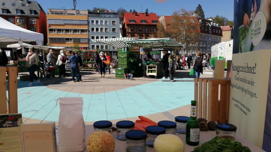 Blick aus einem Infostand auf den Marktplatz mit Gemüseständen und Personen
